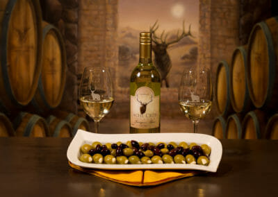 A bottle of Cache Creek Sauvignon Blanc, two filled wine glasses, and a beautifully presented dish of garlic-stuffed green olives and Calamata olives